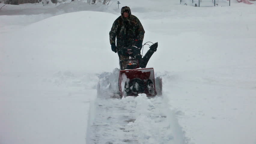 Man pushes a snow blower towards view in deep snow blizzard on sidewalk. Snow blowing in strong wind in sub freezing weather. Winter time hazard and climate nature.
