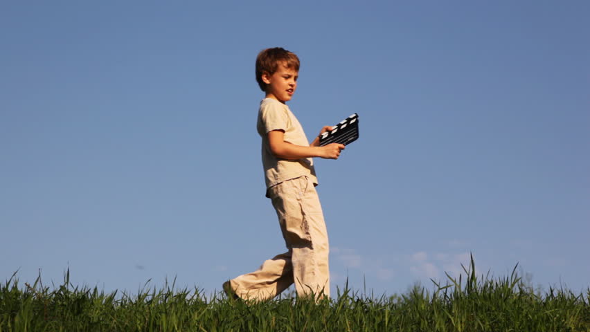 boy walks on grass, says something, clicks clapperboard and goes away against blue sky