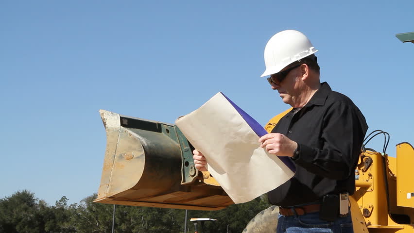 Building inspector in hardhat looks at blueprints while standing in front of a bulldozer at a construction site.