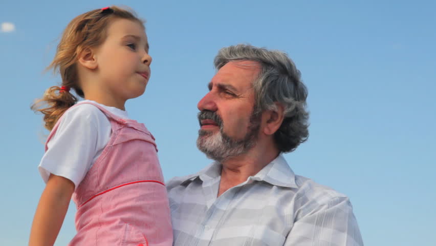 elderly man with his granddaughter on hands, she says something and gesturing against sky in summer
