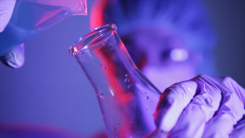 The gloved hand of a technician or scientist reaches down and picks up a test tube with a dark liquid inside and adds to it from a dropper containing a lighter colored substance.