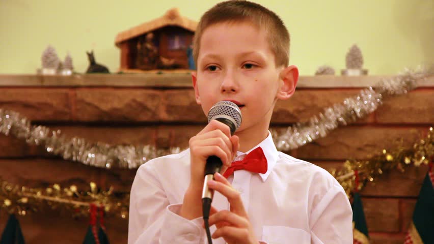 boy in glasses sings Christmas song into microphone in front of fireplace
