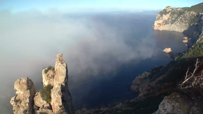 timelapse of a foggy & rugged cliff over the sea, wiht some trees on the right, a big rock boulder on the left