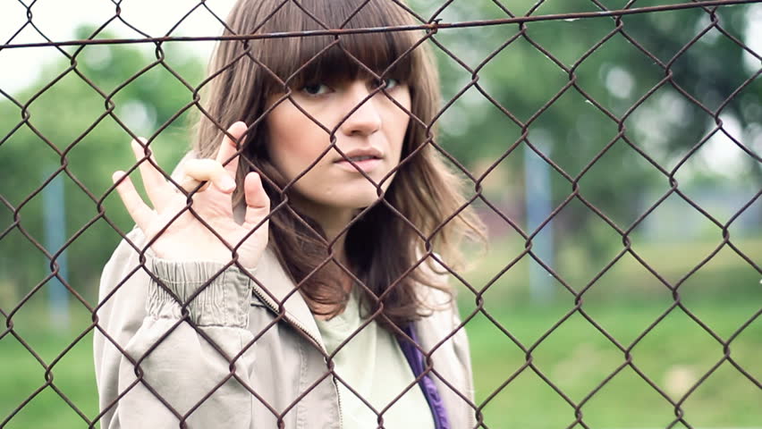 Sad woman behind chain-link fence, outdoors, slow motion
