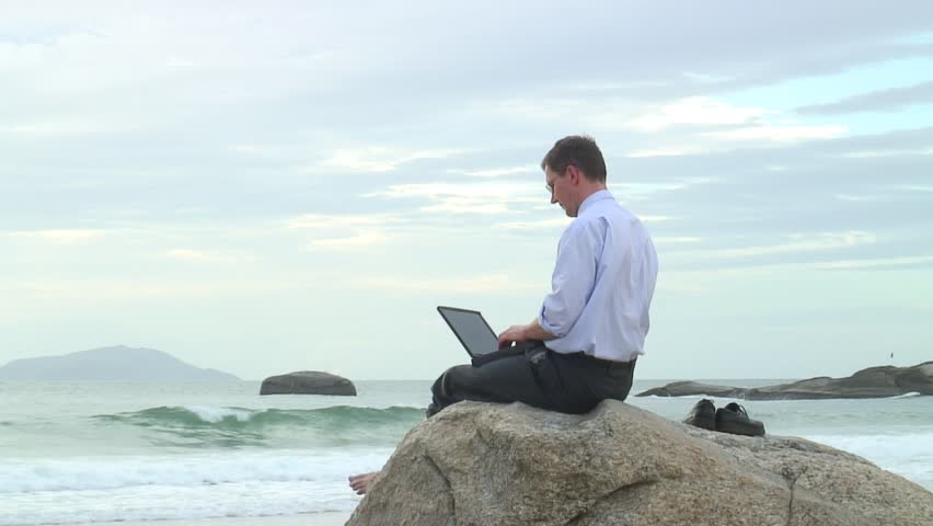 Businessman working with laptop while sitting on a rock on the beach.
