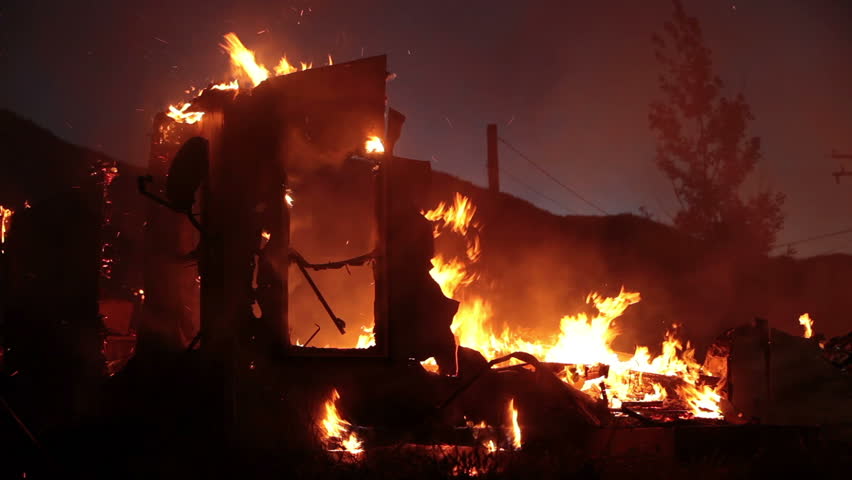 Fireman working and walking as a silhouette on a house fire after dark at night, pushing burning wall down. Flames consume entire home.