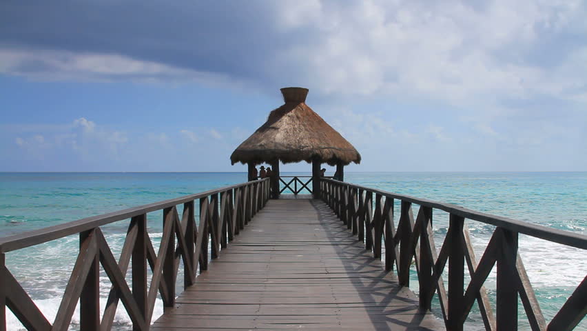 A point of view shot of an ocean pier jutting into the Gulf of Mexico in Cancun, Mexico with people watching the ocean.