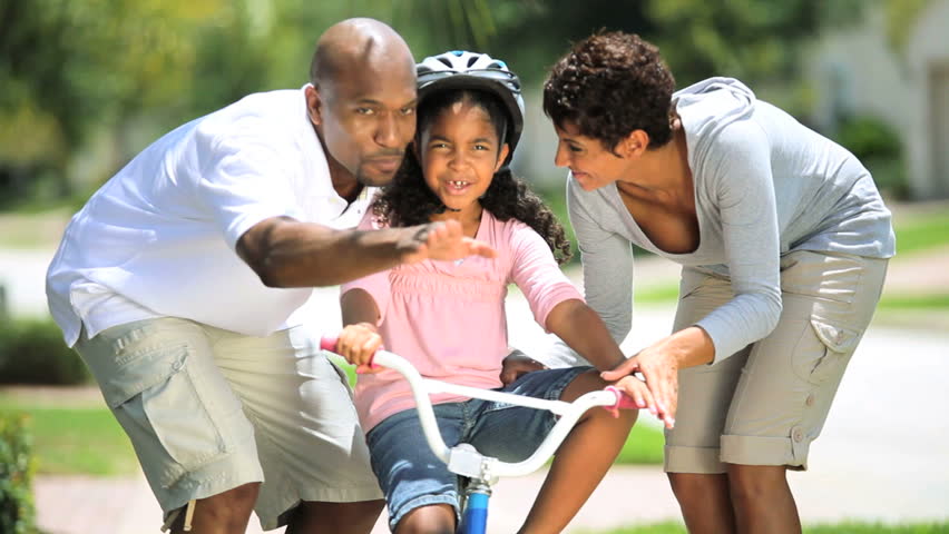 Cute little african american girl being encouraged by her parents as she practices riding her bicycle