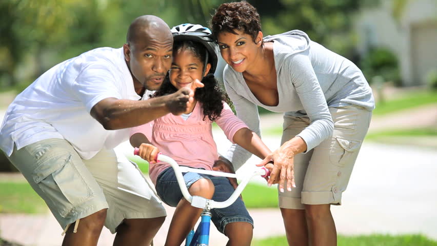 Little african american girl being helped to ride a bicycle by her parents