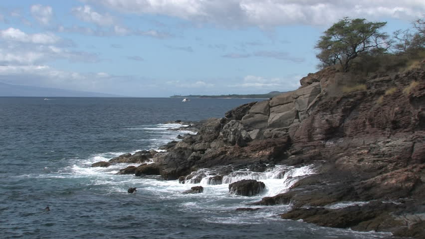 Rocky shoreline on island of Maui in Hawaii. A man  swimming and spear fishing in the rough water. Beautiful water with cliff and trees overlooking water. Island of Lanai on horizon.