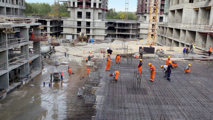 Many builders in action at construction site, machine moves among cranes and unfinished buildings