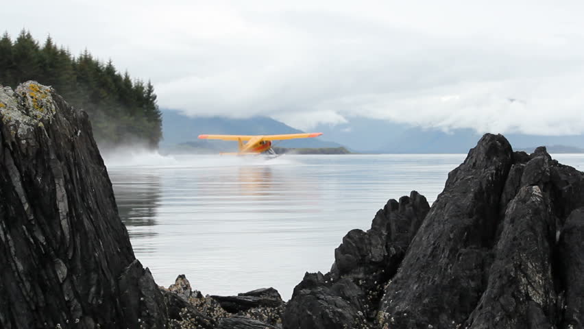Seaplane, framed by beautiful, craggy boulders, glides on rippling water as it prepares to takeoff into thick clouds hanging over mountains. 