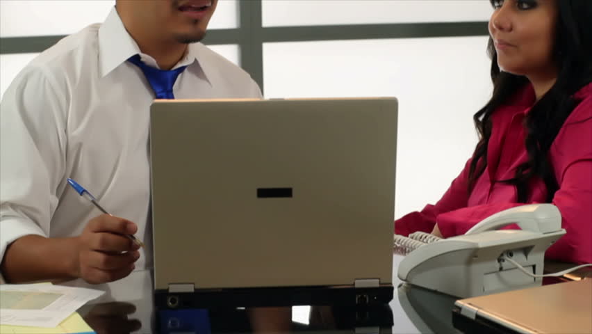 Two attractive Hispanic coworkers engage in conversation about what they see on the laptop, then look up and smile at the camera.