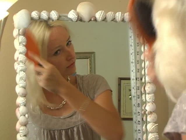 A beautiful blonde brushes her hair in front of a full-length mirror.