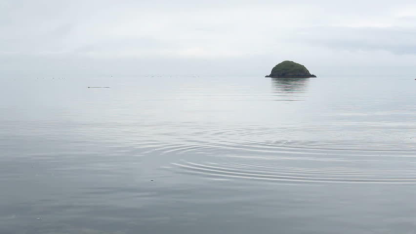 Flat, smooth stone tossed into water ripples across surface towards green, jutting boulder off coast of Kodiak Island, Alaska. 1080p