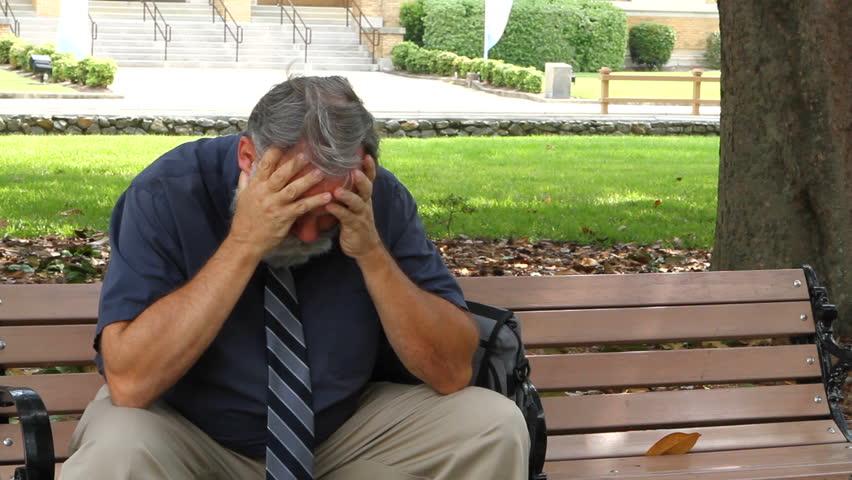 Salesman sits depressed and unemployed on a park bench with his head in his hands.