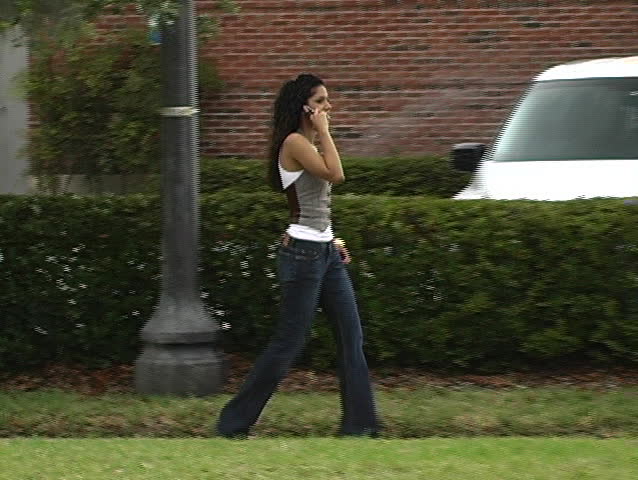 A beautiful Latina brunette walking on a sidewalk, talking on her cell phone.  Full-length.