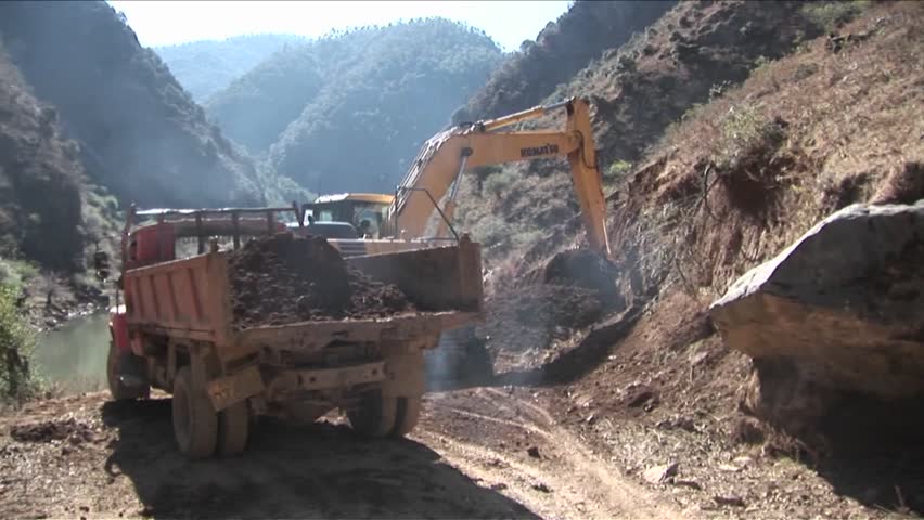 A steam shovel moves earth into a dump truck along a rural road