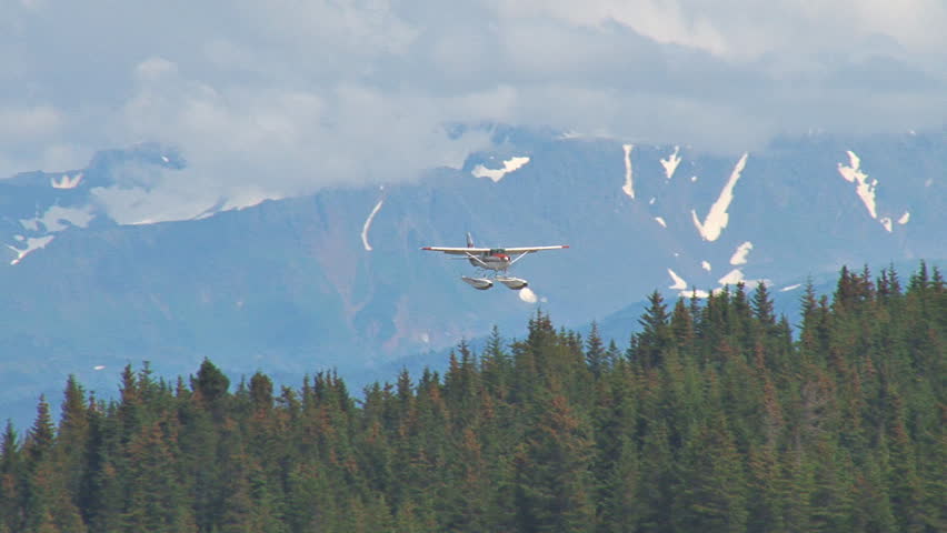 Descending from a cloudy sky with mountains in the background, a red and white floatplane lands on a lake with houses and other planes in the background.