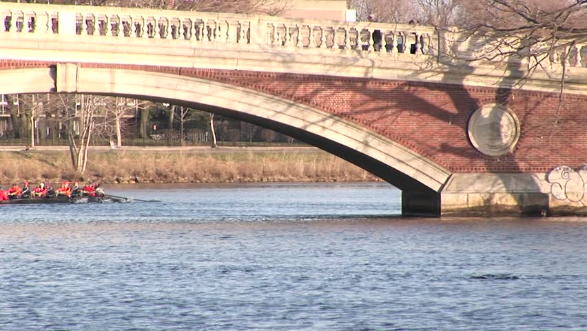 Long-shot of the Harvard University rowing team passing under a bridge over the Charles Rives near Cambridge, Massachusetts.