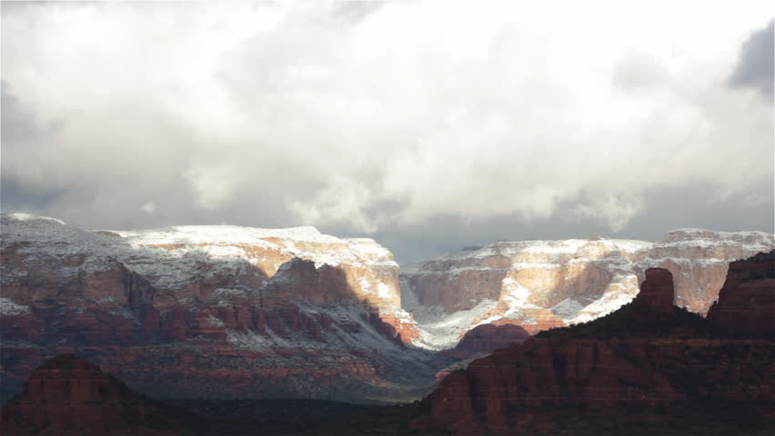 Sunlight shines down through heavy, early-morning clouds covering a mountainous canyon near Sedona, Arizona with a light dusting of snow. Wide shot.