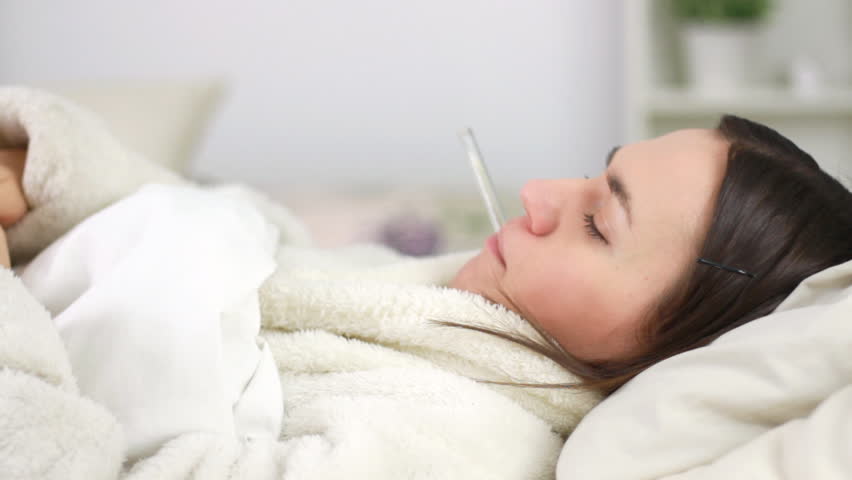 Sick woman in bed checking her temperature and blowing nose
