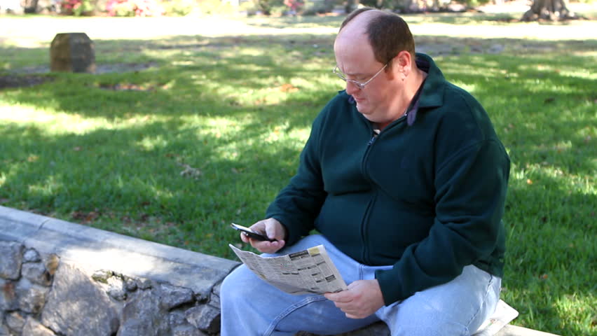 Unemployed obese man uses a cell phone to call about a job in the want ads section of the newspaper while in the park.