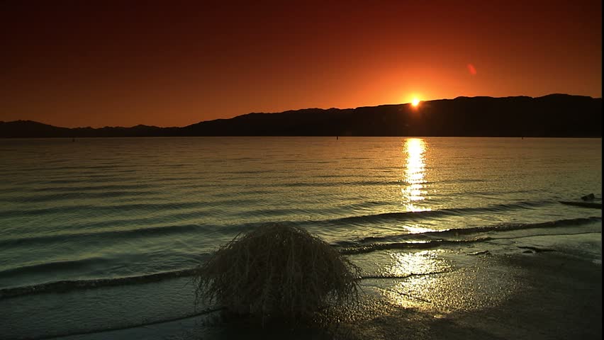Dolly left as sun rises over lake Mead, with tumbleweed in foreground at edge of water