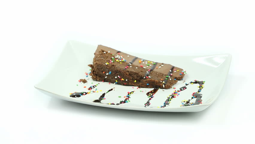 Tasty piece of chocolate cake on a large white plate, covered in chocolate syrup and colorful sprinkles getting picked up and placed back on white table surface.