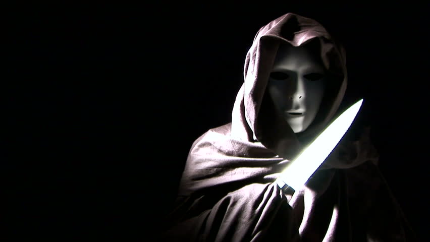 A person masked and cloaked in black, flashing a knife.