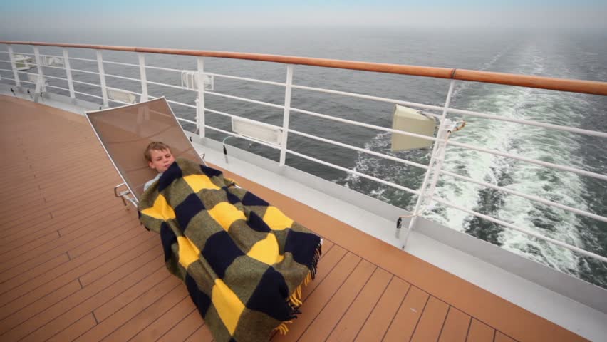 Little boy lies under plaid on deckchair at deck of ship during cruise