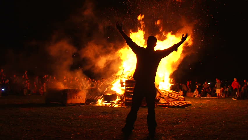 Man in front of large bonfire has both arms up high taking in the heat, filmed in slow motion.