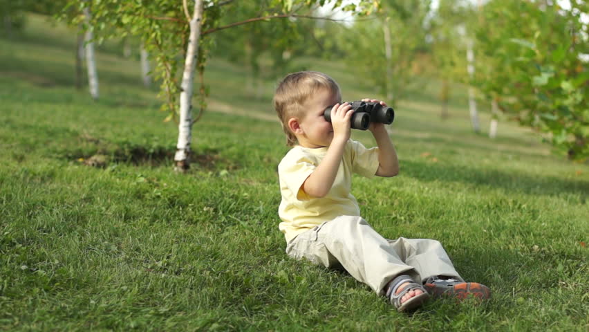 Little boy with binoculars sitting on the grass. Thumbs up. Ok.
