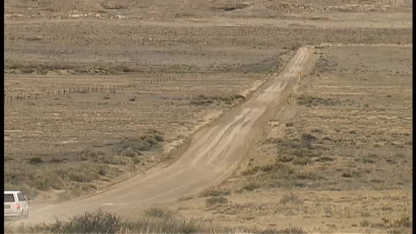 A white minivan moves slowly and carefully down a rough dirt road in the New Mexico desert, going away from the camera