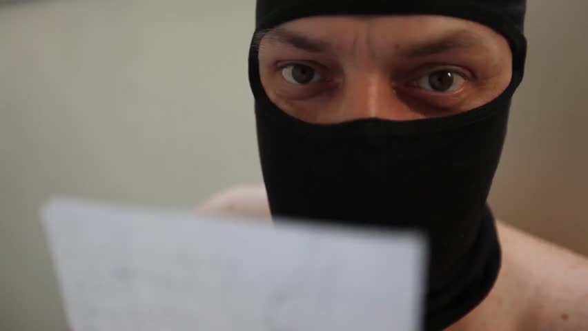 Naked young adult man with black balaclava reads a paper.

