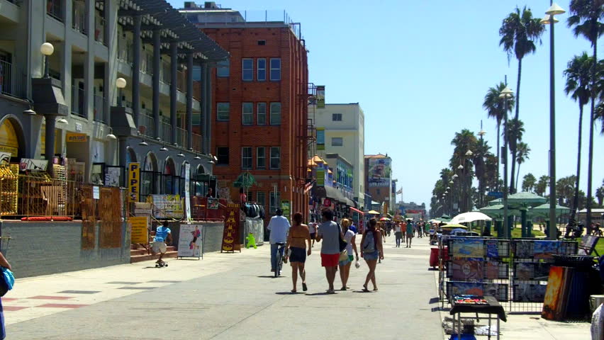 A shot looking down the Venice Beach Boardwalk with souvenir shops, historic buildings, palm trees and people walking. 
