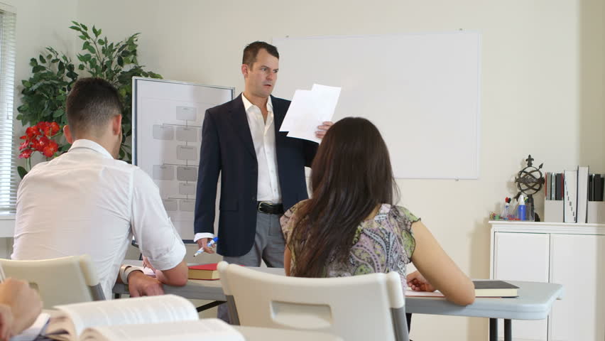 Adult students in a classroom setting listen attentively to their instructor working at the whiteboard.