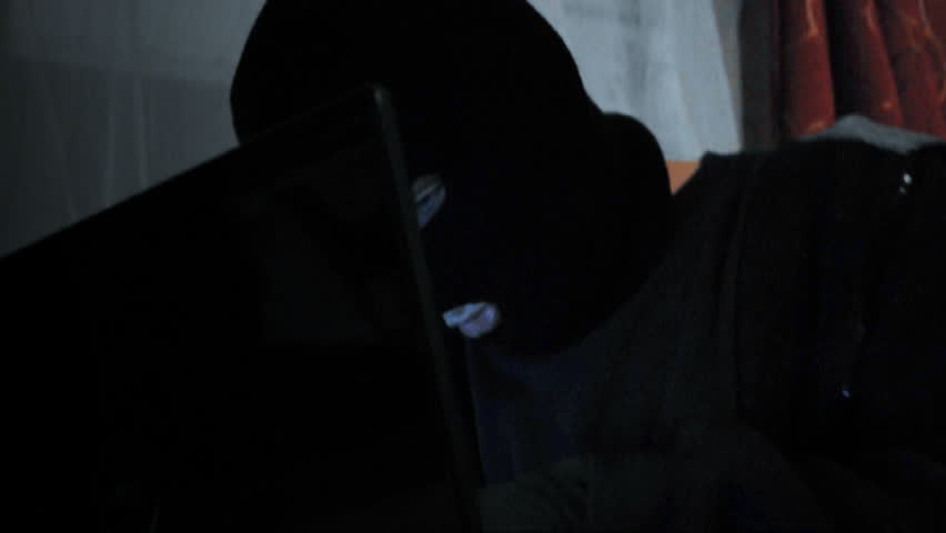 A computer hacker is breaking into a laptop and reading secret information. He is wearing a black mask.