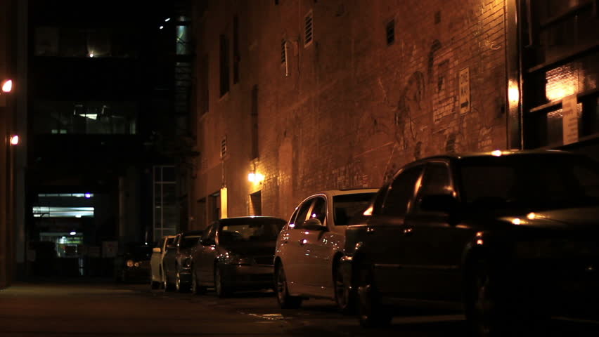 A suspicious man in a hooded sweatshirt walks away down a dark alley downtown