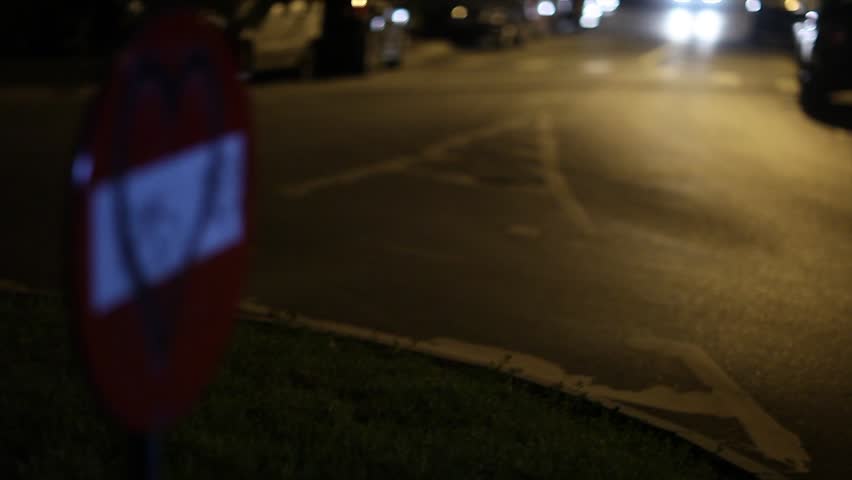 Blonde girl walking on the street at night, passing and slowly caressing a âNo Entryâ traffic sign with a graffiti heart on it, while a taxi passes in the background.
