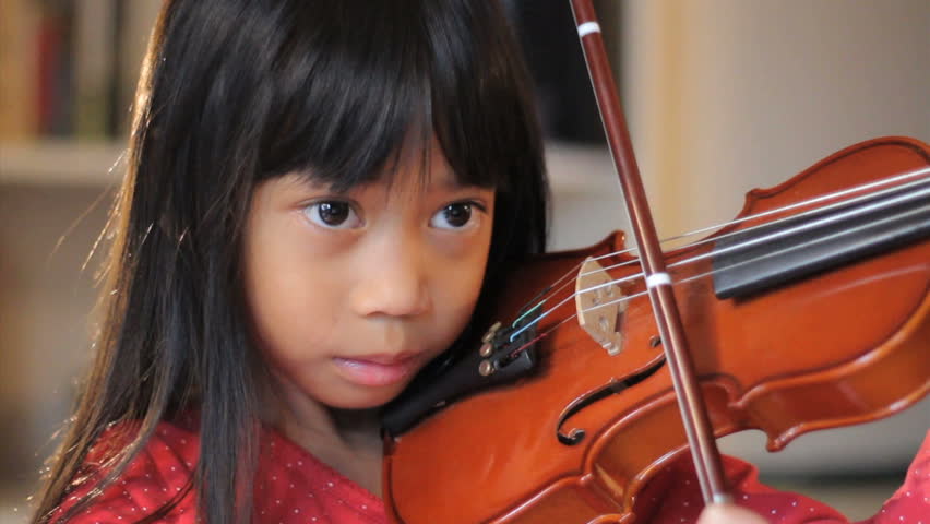 A close up shot of a pretty 6 year-old Asian girl diligently practices her violin in the living room.
