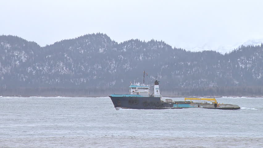 KACHEMAK, AK - CIRCA 2012: An impressive and imposing heavy-duty tug/utility vessel returns to the calmer bay, passing a rolling container barge being towed by a smaller tug.