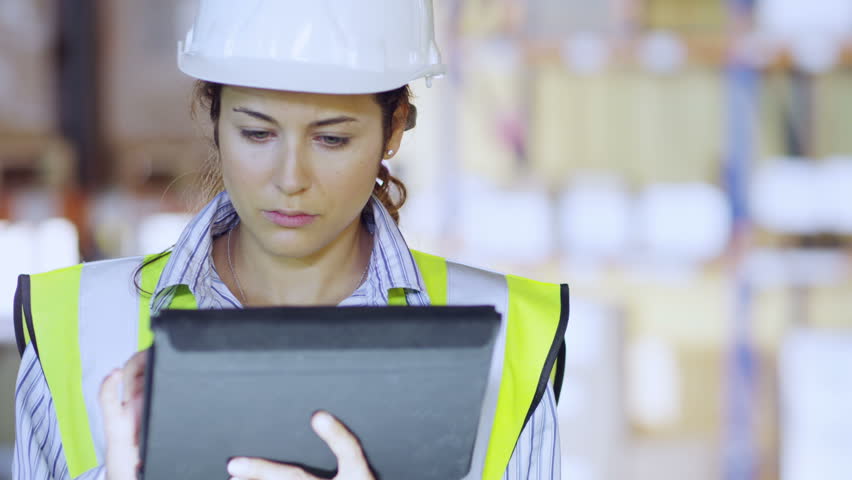 An attractive female warehouse employee wearing high visibility clothing and a hard hat is working on a digital tablet and checking stock levels. Other employees can be seen working in the background
