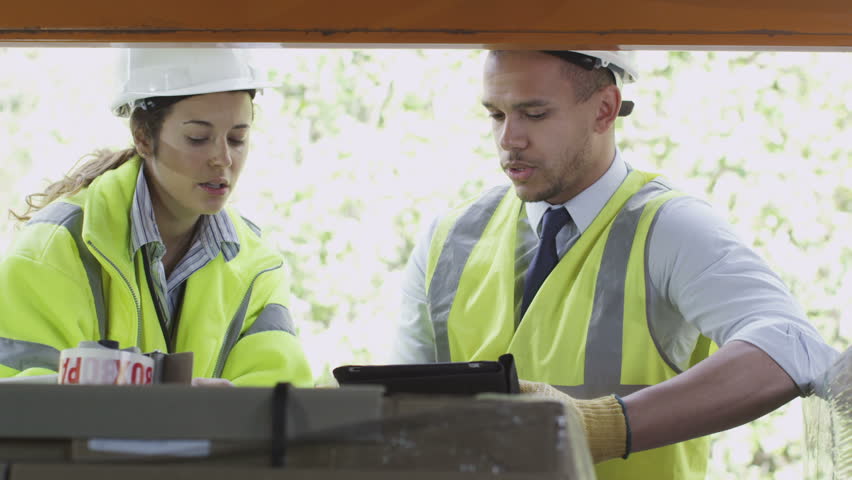 male and female warehouse workers in hard hats and hi visibility clothing chatting together while they work