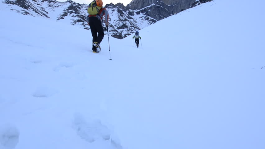 Two climbers hike up a snow covered hill towards a mountain peak covered in clouds during the day in Alaska