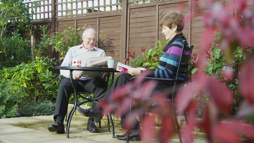A mature couple of retirement age are spending time together sitting in their garden. They are chatting and drinking cups of tea and reading magazines.