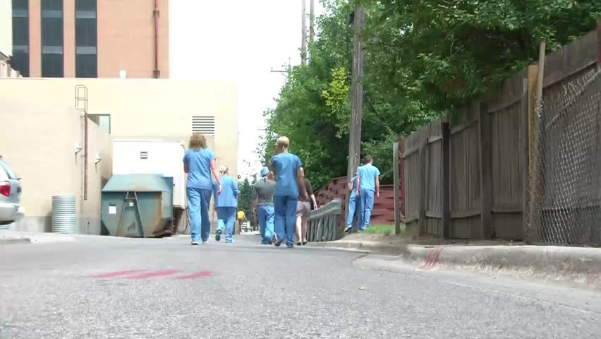 Hospital staff workers walk away from camera in mass in blue health care attire with doctor in white coat towards end.