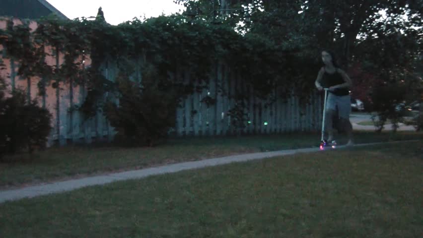 Girl on lighted scooter passes by on sidewalk as dusk turns into night with wheels illuminating yielding colorful effect.