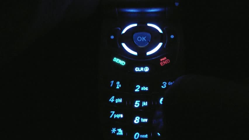 Illuminated cordless cell phone displays colorful lights in the dark while dialing numbers with thumb.