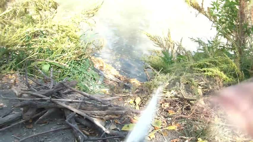 Survival clip of knife carving stick into sharp point on river bank with a pile of sticks for fire in frame.